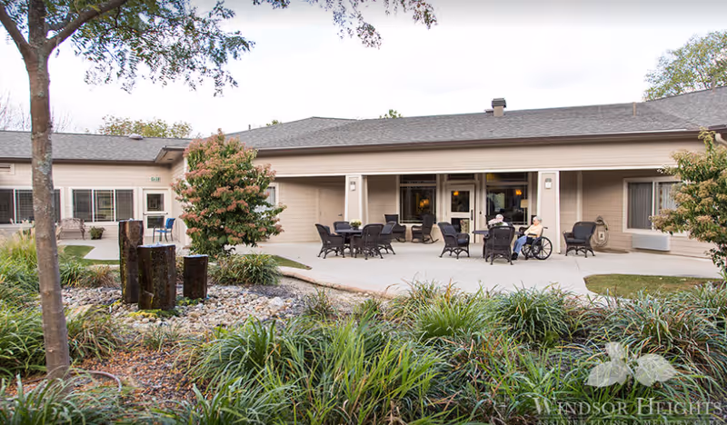 Outdoor patio area of Windsor Heights Assisted Living and Memory Care with several black wicker chairs and tables arranged on a concrete surface. Two elderly individuals are seated, one in a wheelchair. The patio is surrounded by greenery, including bushes, grass, and trees. The building exterior is beige with large windows and a covered walkway.