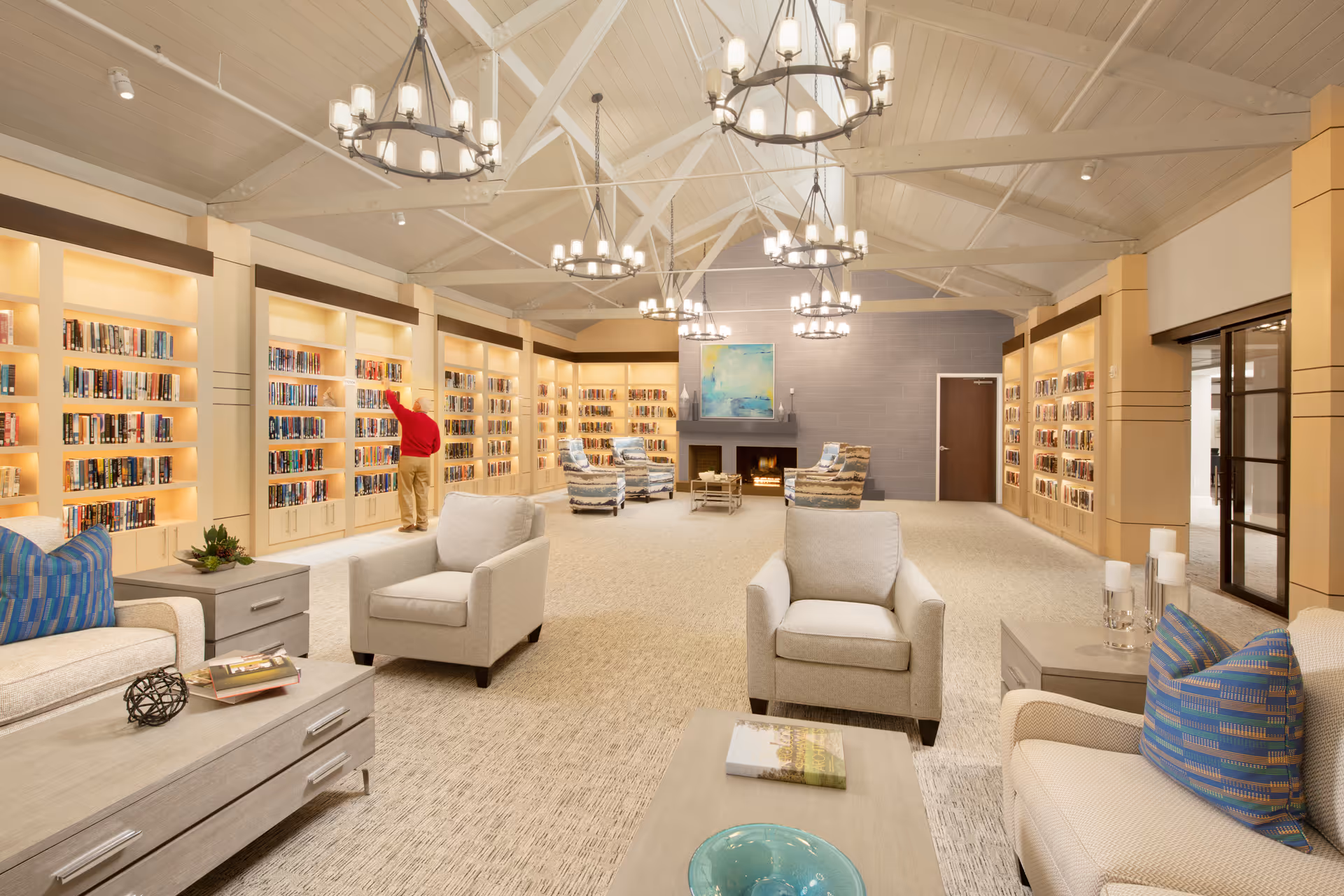 A spacious and well-lit library room with beige carpet and light wood-paneled walls. The room features multiple built-in bookshelves filled with books, several comfortable armchairs and sofas with blue patterned pillows, coffee tables, and modern chandeliers hanging from a vaulted ceiling. A person in a red sweater is selecting a book from one of the shelves. There is a fireplace with a painting above it at the far end of the room.