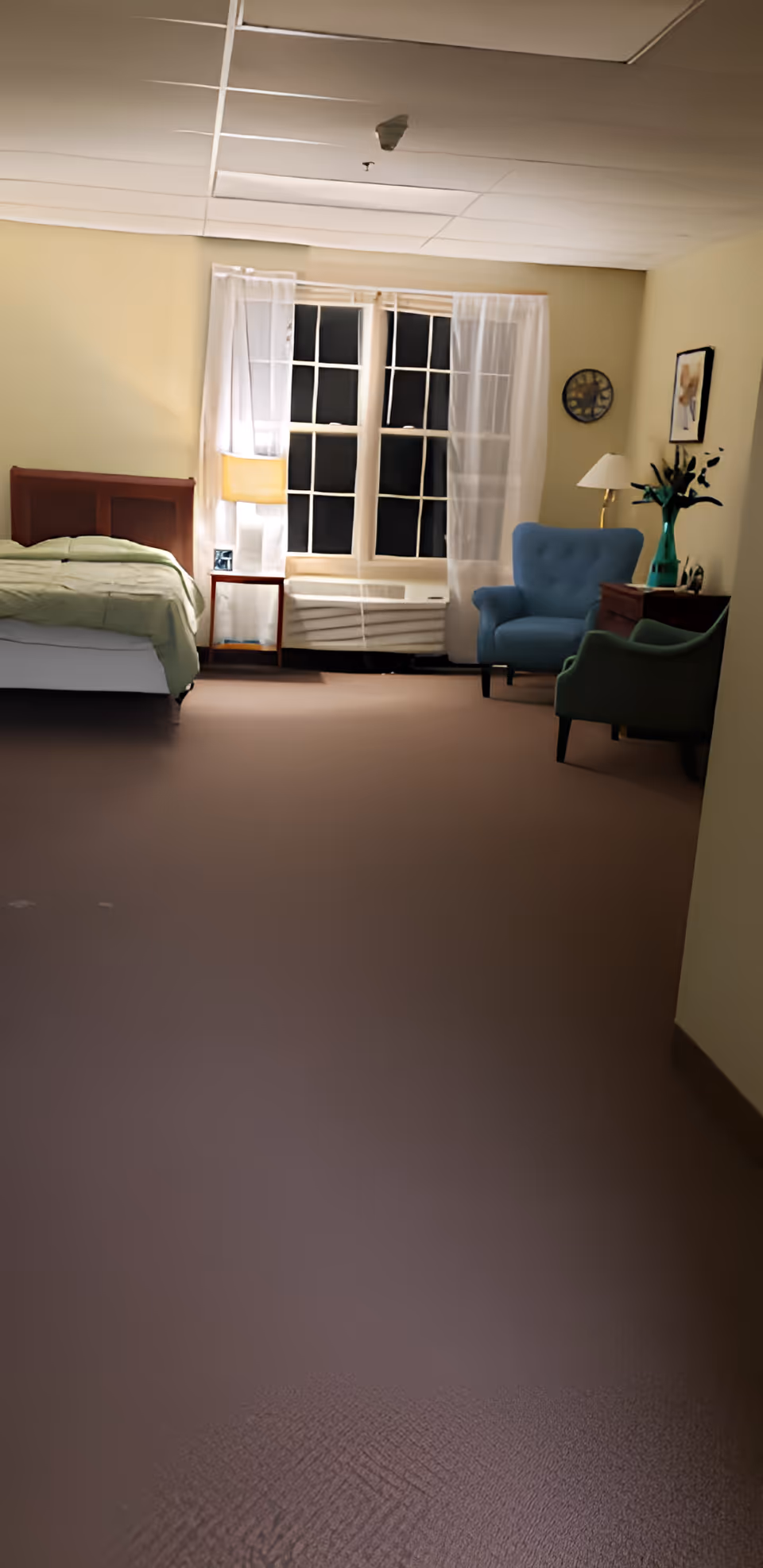 A senior living bedroom with a single bed, a window with sheer curtains, a bedside lamp, and two upholstered chairs.