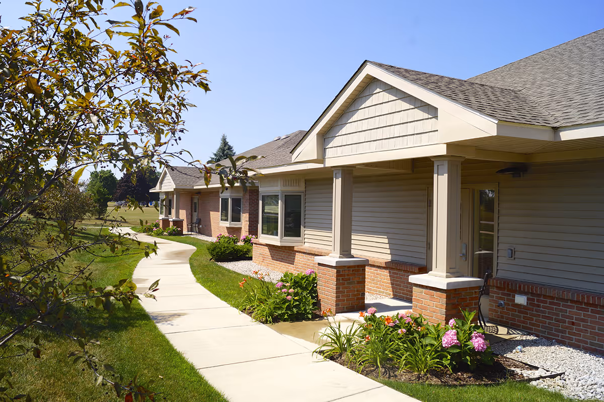 Sidewalk leading past a single-story senior living building with a covered porch, columns, and landscaped flower beds.
