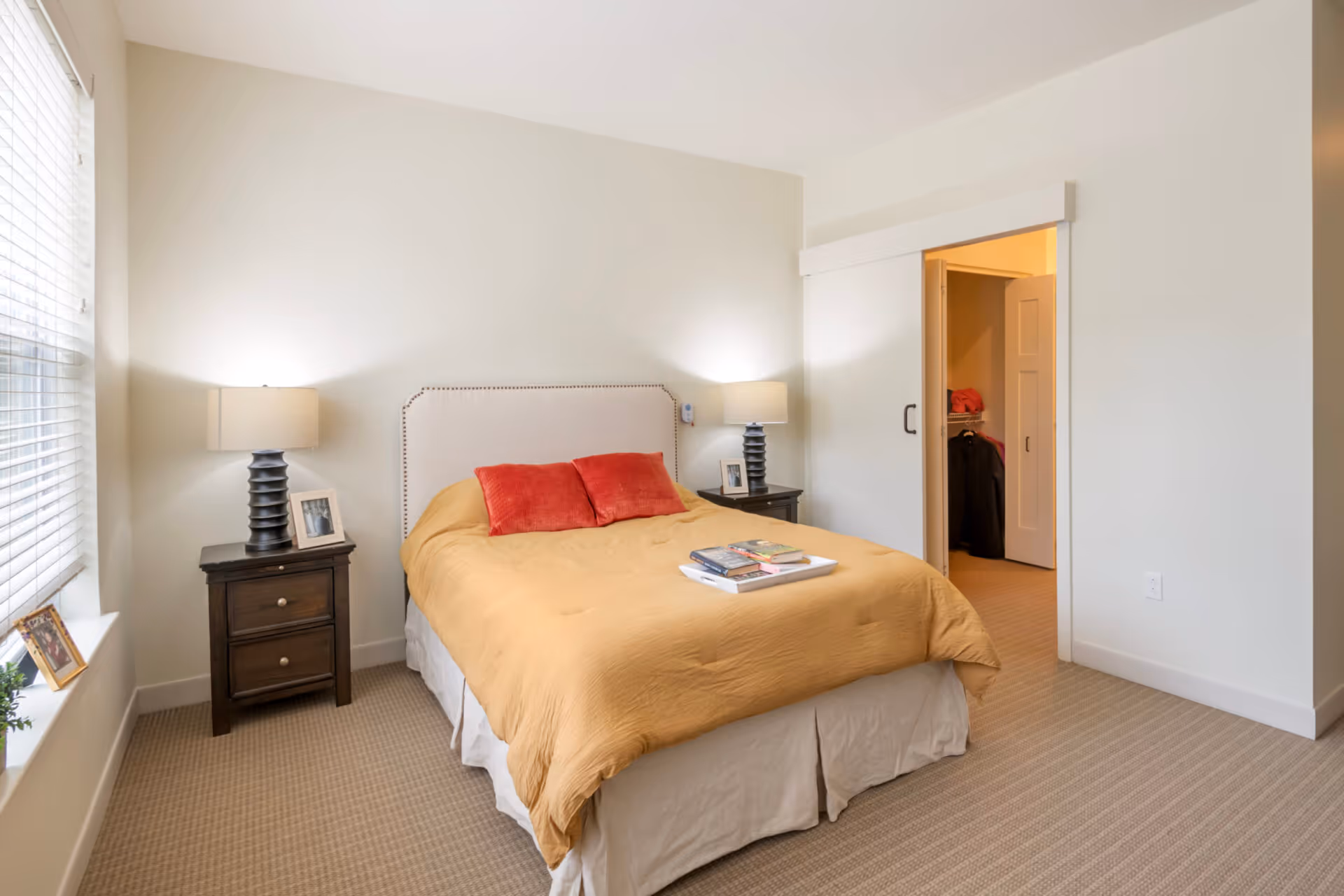 A bedroom with a bed covered in a mustard yellow bedspread and two red pillows. There are two dark wooden nightstands on either side of the bed, each with a lamp and a framed photo. A window with blinds is on the left side of the room, and an open sliding door reveals a walk-in closet with clothes inside. The walls are painted white and the floor is carpeted.
