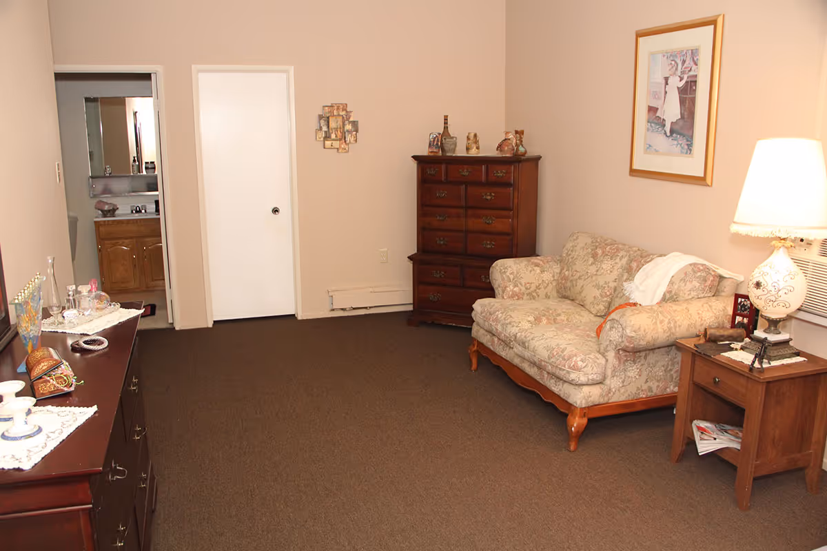 A cozy living room with beige walls and brown carpet. There is a floral upholstered loveseat next to a wooden side table with a decorative lamp and some magazines underneath. On the opposite side, there is a wooden dresser with various decorative items on top. A framed picture hangs on the wall above the loveseat. Two doors are visible, one leading to a bathroom with a sink and mirror, and the other is closed. A small photo collage is mounted on the wall between the doors.