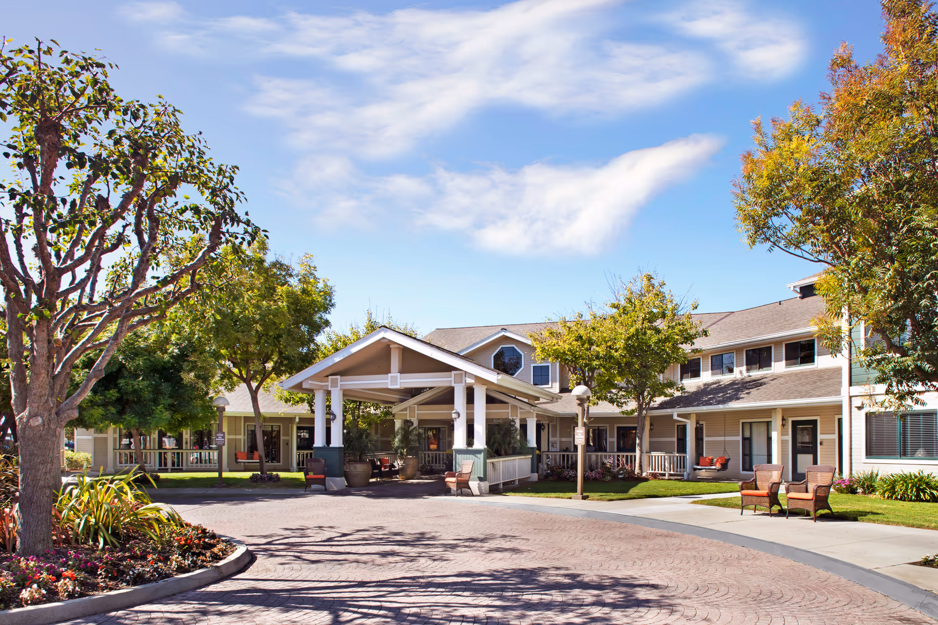 Front entrance of a two-story senior living building with a covered porte-cochère, trees, and outdoor seating.
