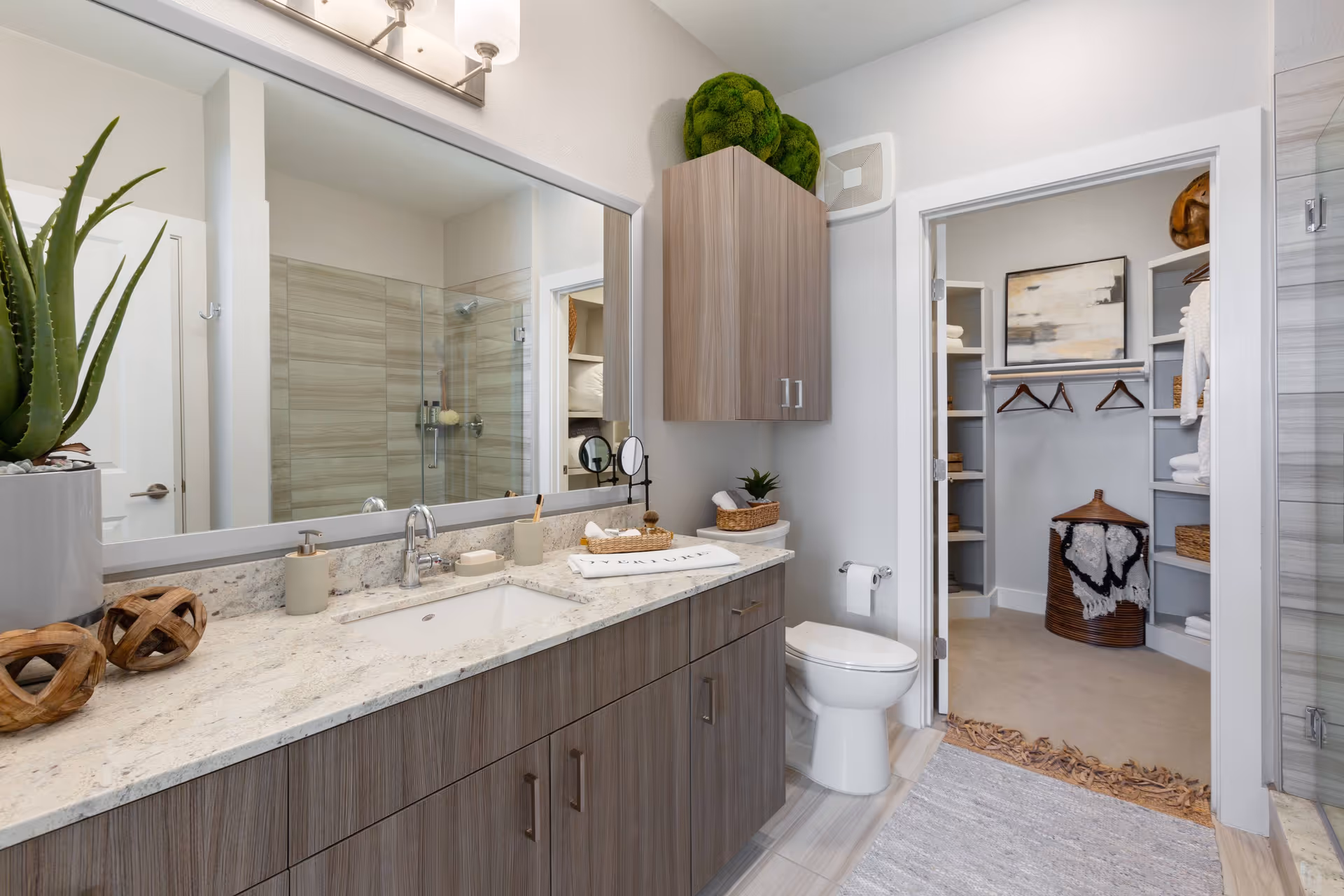 Modern bathroom with a long vanity and sink, large mirror, toilet, glass-enclosed shower, and a doorway leading to a walk-in closet.