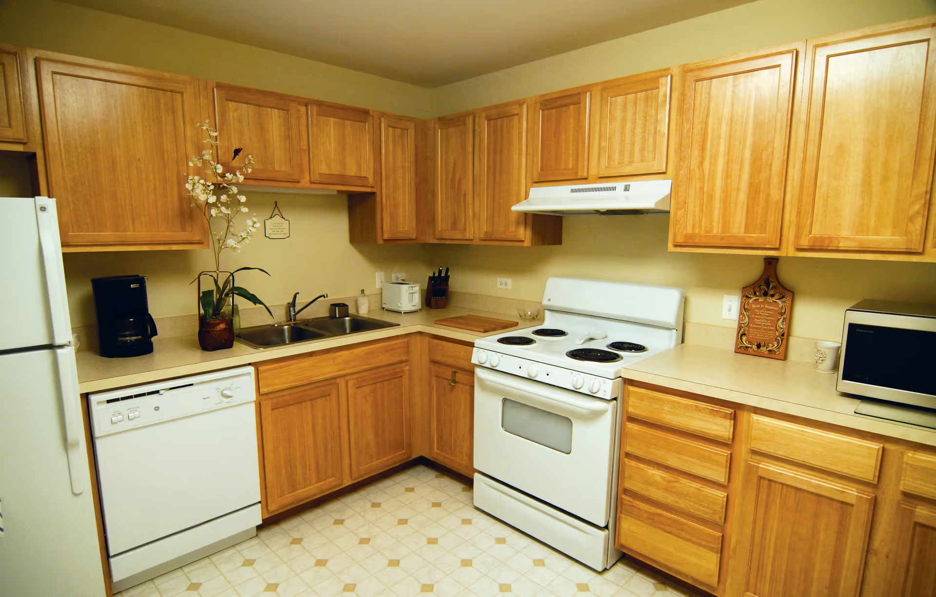 A kitchen with wooden cabinets, a white refrigerator, dishwasher, electric stove with oven, microwave, coffee maker, and a double sink. There is a potted plant with white flowers on the counter and a decorative cutting board on the right side.