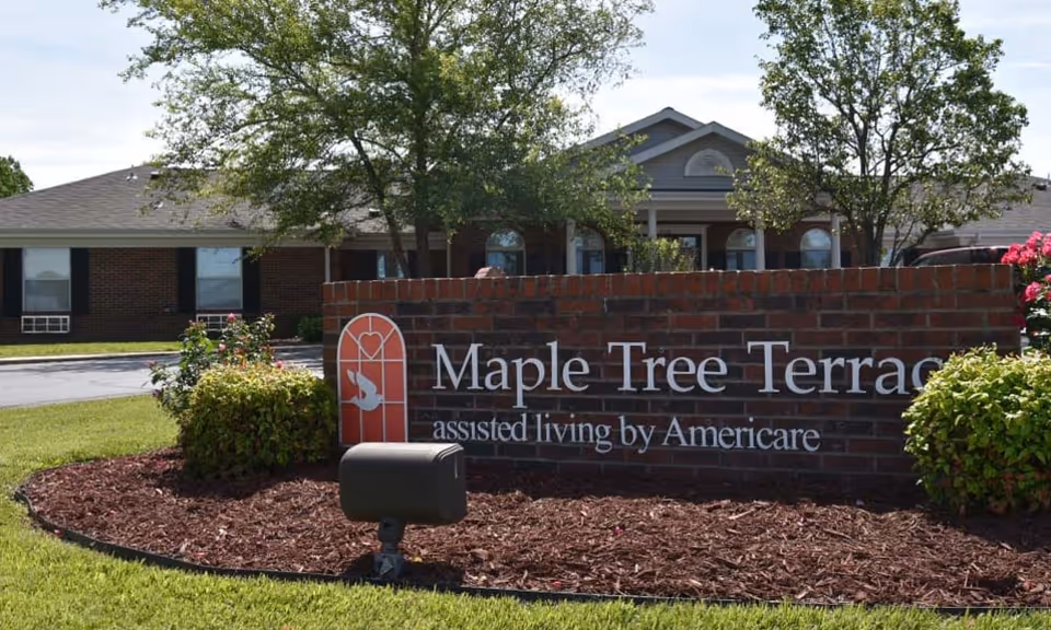 Brick sign for Maple Tree Terrace assisted living by Americare in front of a single-story brick building with trees and landscaping around.
