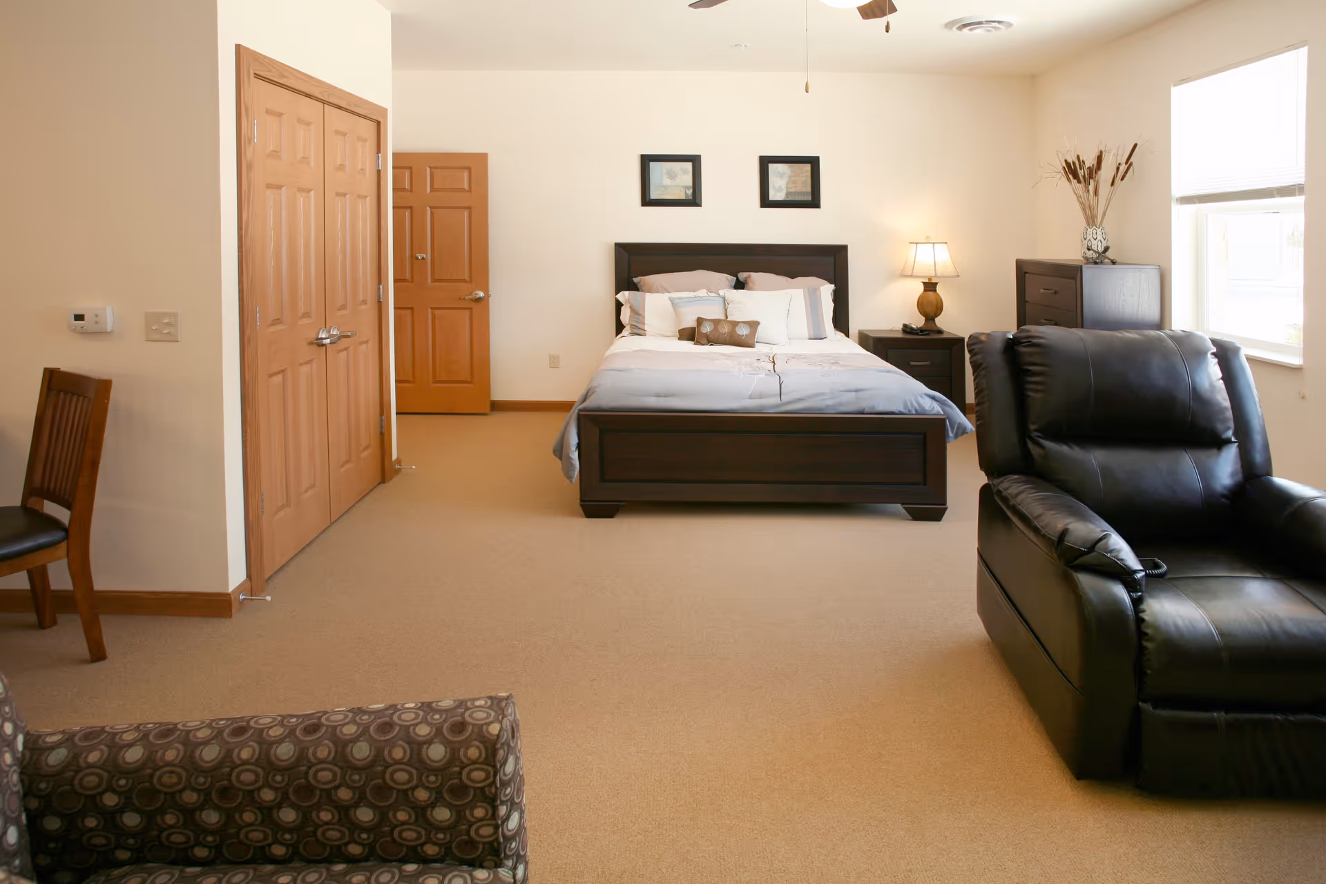 A spacious bedroom with a large bed centered against the back wall, flanked by two nightstands with a lamp on the right side. There is a wooden chair on the left, a patterned armchair in the foreground, and a black leather recliner on the right near a window. The room has beige carpet, light-colored walls, and wooden doors and trim.