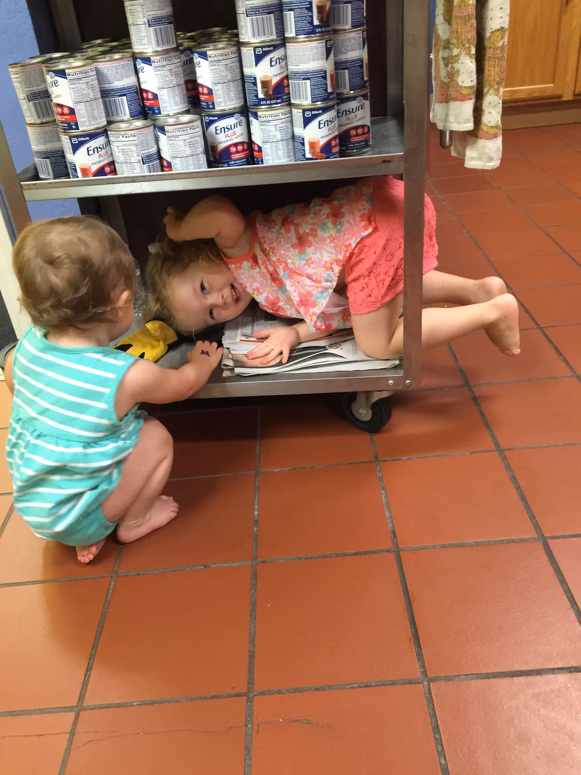 Two toddlers playing around and one lying under a metal cart stocked with Ensure cans on a tiled floor.