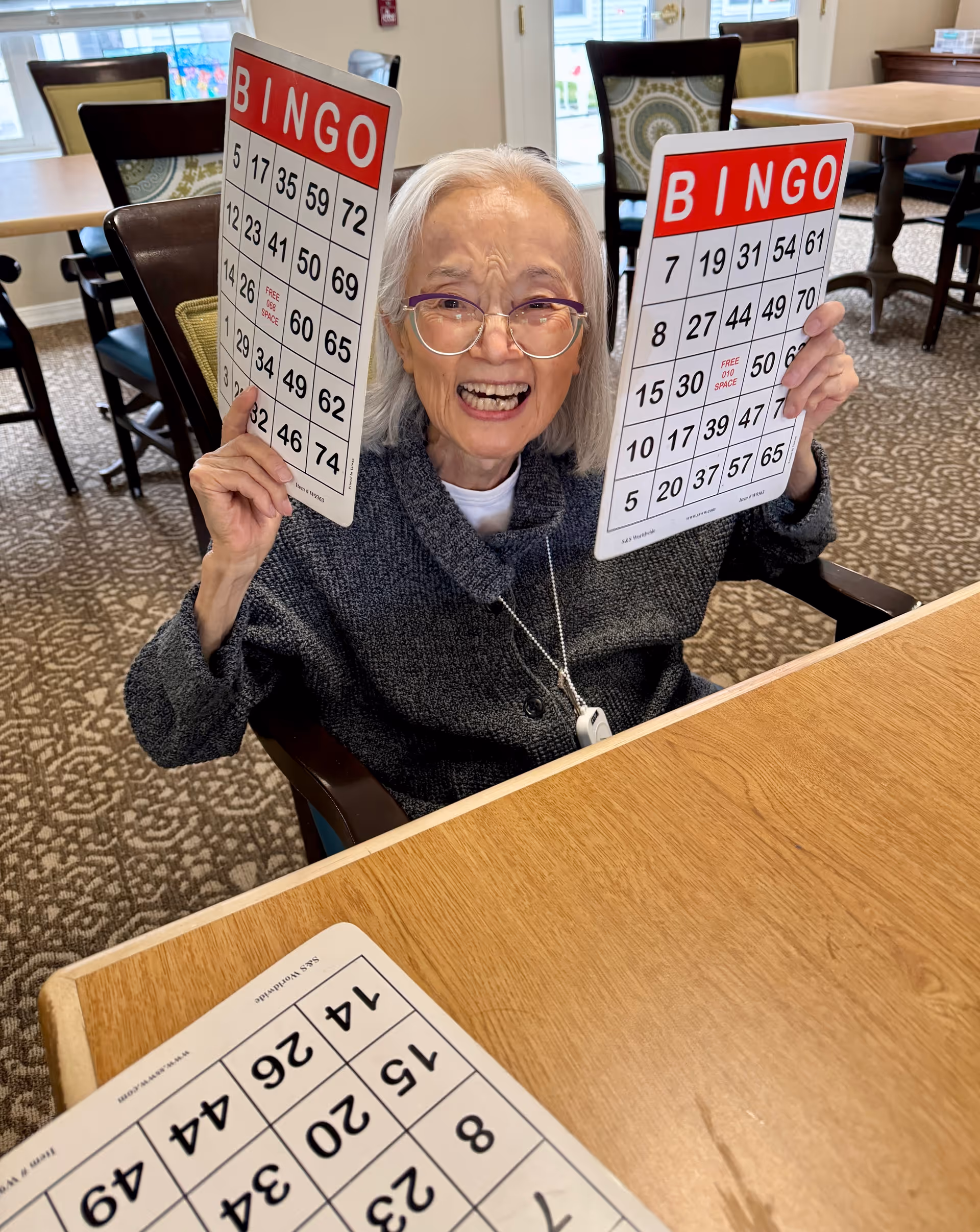 An elderly woman with gray hair and glasses is sitting at a table in a senior living facility, smiling and holding up two bingo cards. The room has patterned carpet, wooden tables, and chairs with cushions, with windows in the background letting in natural light.
