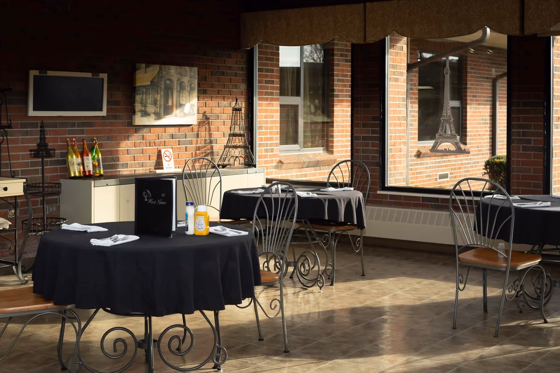 A dining area with round tables covered in black tablecloths, each set with napkins and condiments. The room has large windows showing a brick exterior wall and features decorative Eiffel Tower models and a painting on the brick wall inside.