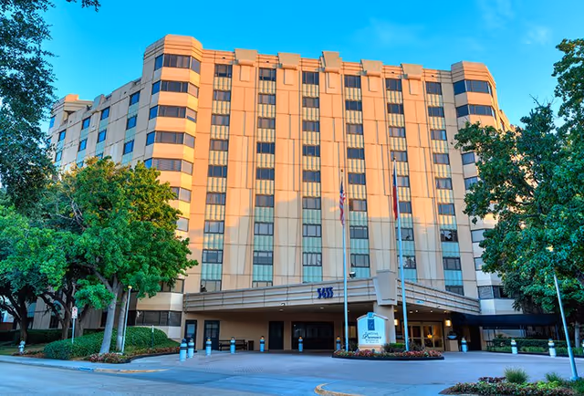 Front exterior view of a multi-story beige building with many windows, surrounded by green trees and landscaping, with a driveway and entrance canopy. The building is identified as Five Star Premier Residences Of Dallas.