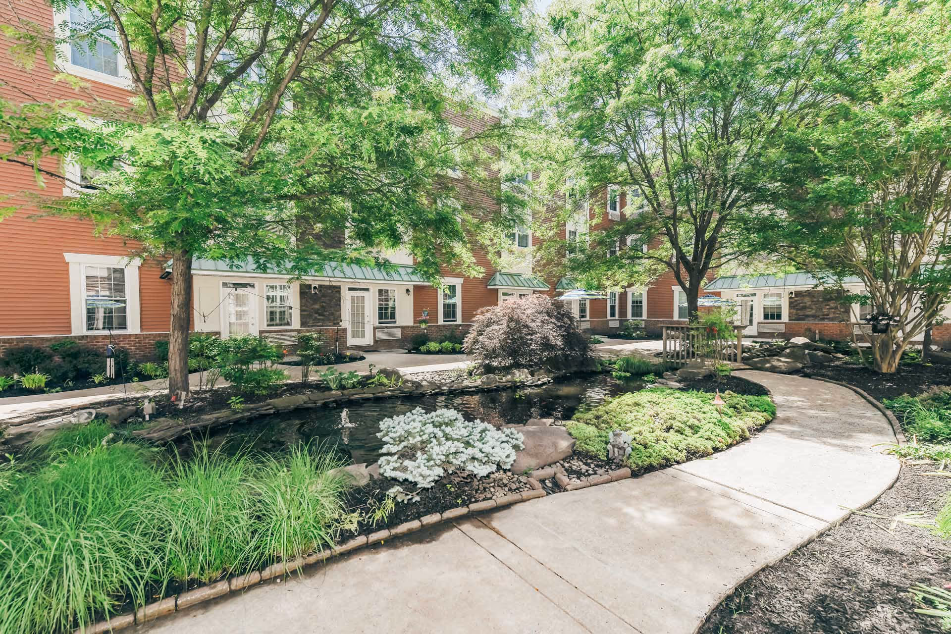 A serene outdoor courtyard at Woodbury Mews featuring a winding concrete pathway, lush green trees, shrubs, and a small pond with rocks and plants surrounding it. The courtyard is enclosed by a multi-story building with red siding and white-trimmed windows and doors.