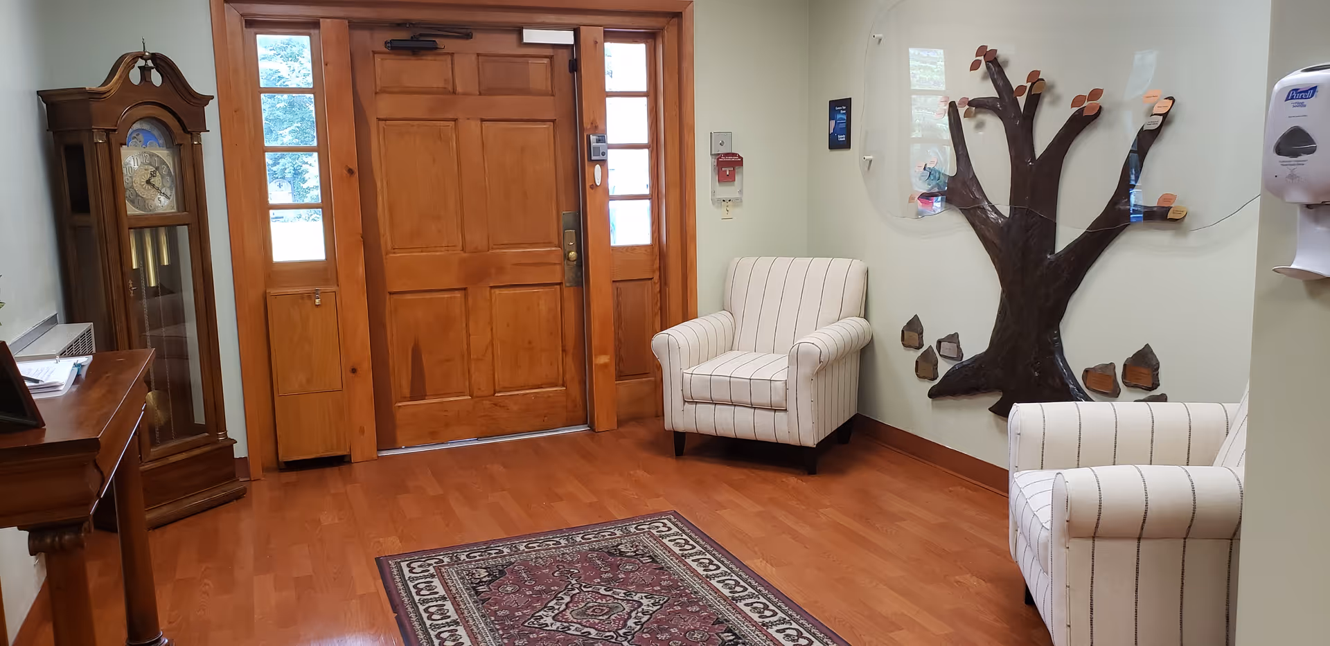 A small seating area near a wooden entrance door with two white armchairs featuring black stripes, a wooden grandfather clock, a wooden table, a decorative wall-mounted tree with small shelves, a patterned rug on a wooden floor, and a wall-mounted hand sanitizer dispenser.