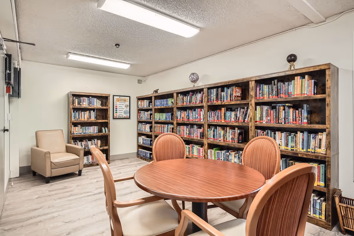 A cozy library room in a senior living facility with wooden bookshelves filled with books along the walls, a round wooden table with four chairs, and a beige armchair in the corner. The room has light-colored walls and flooring with overhead fluorescent lighting.