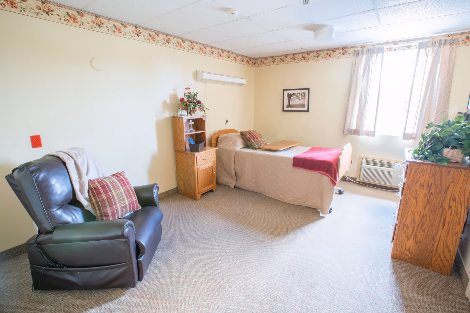 A bright and cozy bedroom in a senior living facility featuring a single bed with beige bedding and a red throw blanket, a wooden nightstand with a floral arrangement, a comfortable dark leather recliner with a plaid pillow and a light blanket, a wooden dresser with a green plant on top, and a window with light curtains letting in natural light.