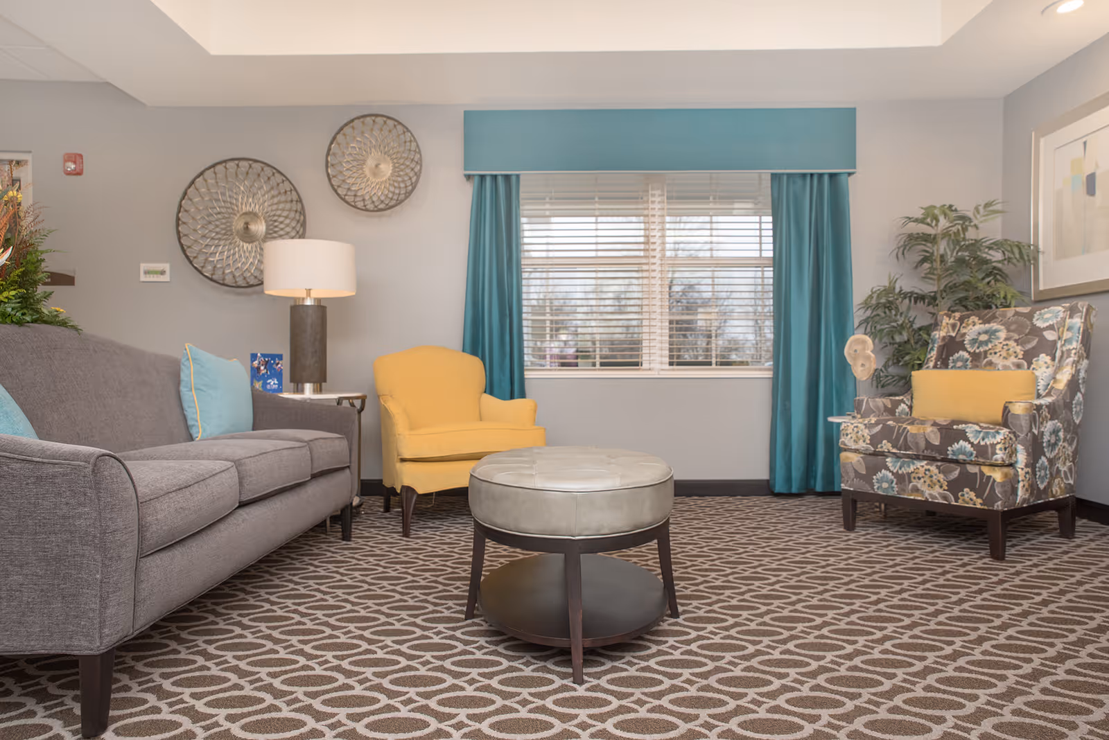 A cozy living room with a gray sofa adorned with blue pillows, a yellow armchair, and a floral-patterned armchair with a yellow cushion. A round ottoman is centered on a patterned carpet. The room features a window with blue curtains, two decorative wall hangings, a floor lamp, a potted plant, and framed artwork on the wall.