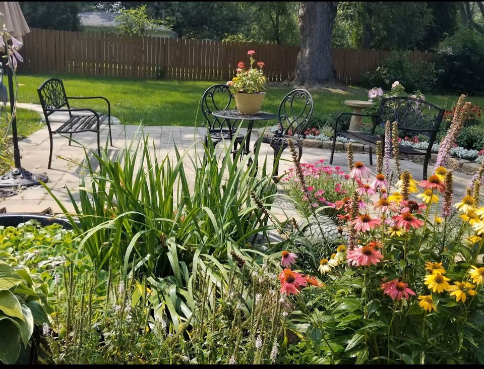 Sunny garden patio with metal benches and chairs, a small table, and colorful flowers in the foreground.