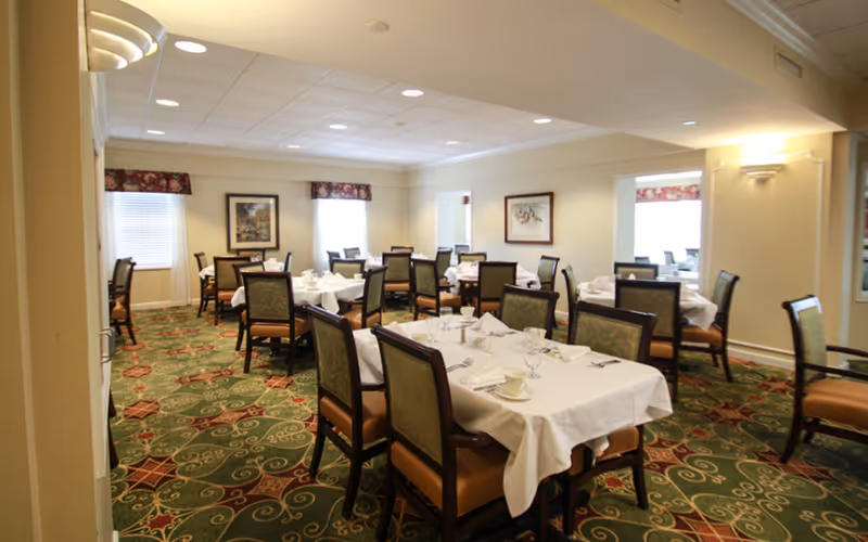 A dining room with multiple tables covered in white tablecloths, set with glassware, cups, and napkins. The room has patterned green carpet, cream-colored walls, framed artwork, and windows with floral valances.