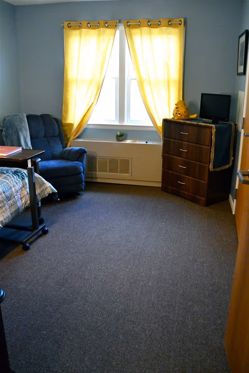A small bedroom with a blue recliner chair next to a bed with a plaid blanket. There is a wooden dresser with a small TV and a decorative yellow bird on top. The room has a window with bright yellow curtains letting in natural light, and the floor is carpeted in a dark color.