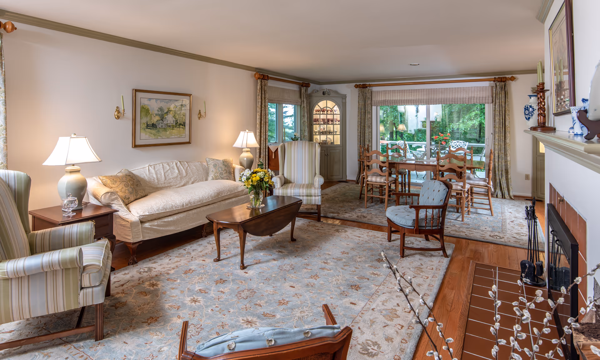 Bright traditional living room with a sofa, armchairs, coffee table and rug, opening to a dining area with a table and chairs and large windows looking out to greenery.