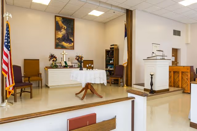 Interior of a small chapel or worship area with a raised platform featuring a white altar table covered with a lace cloth, a lectern, chairs, an American flag, a Christian flag, a piano, and religious decorations including a cross and a painting on the wall.
