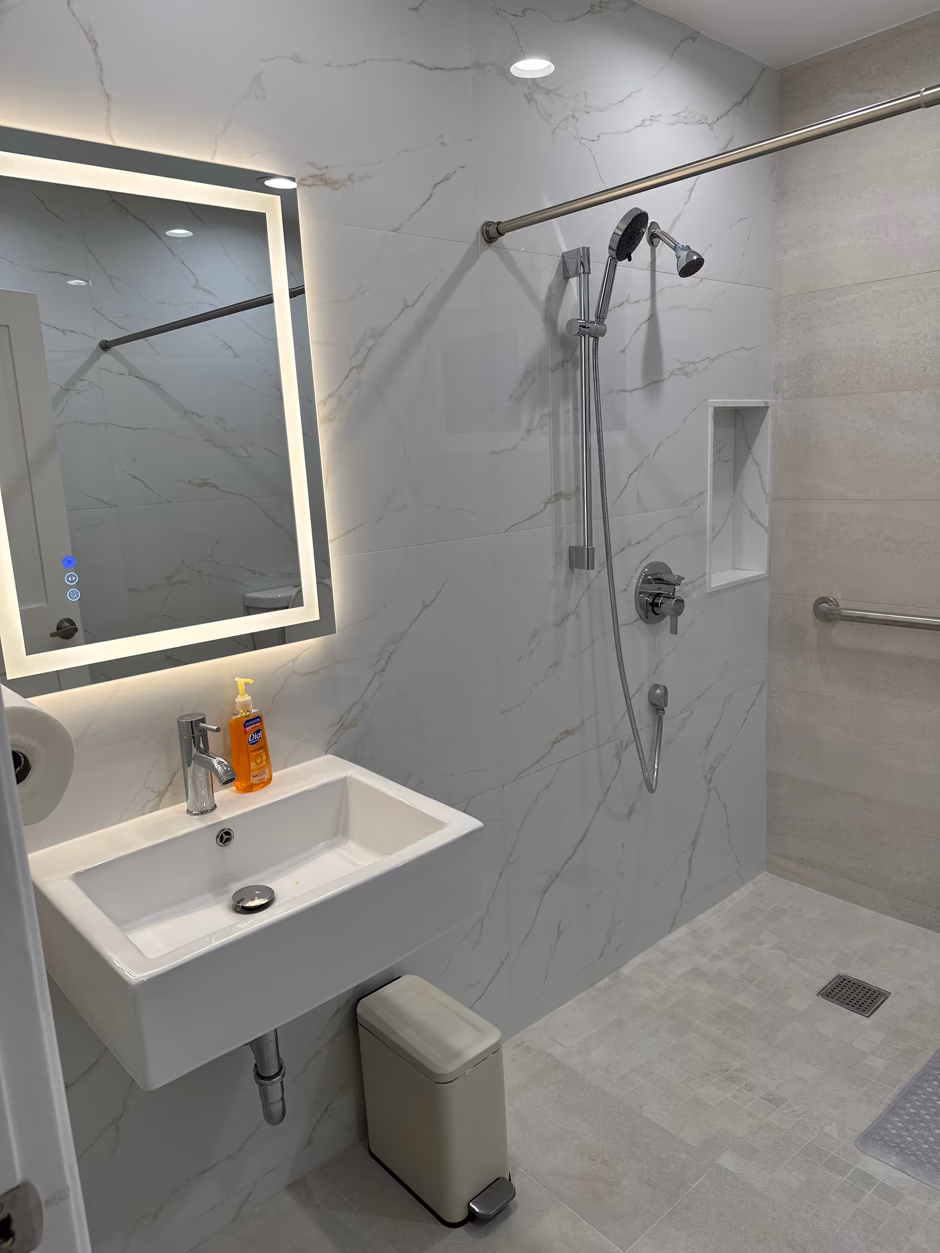 A modern bathroom with white marble-patterned walls and light gray tiled floor. It features a rectangular white sink with a chrome faucet, a backlit rectangular mirror above the sink, a wall-mounted soap dispenser with orange soap, a small beige trash bin below the sink, and a walk-in shower area with a handheld showerhead, a built-in wall niche, and a horizontal grab bar.
