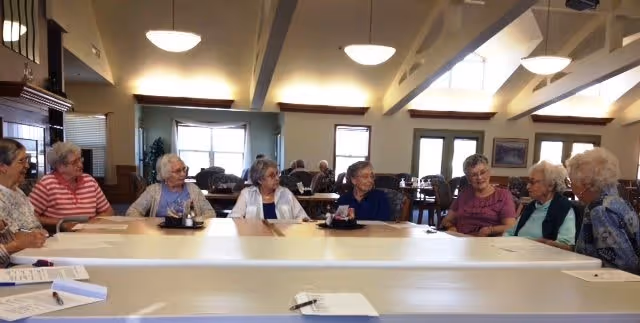 A group of elderly women sitting around a large table in a well-lit room with high ceilings and windows, engaged in conversation.