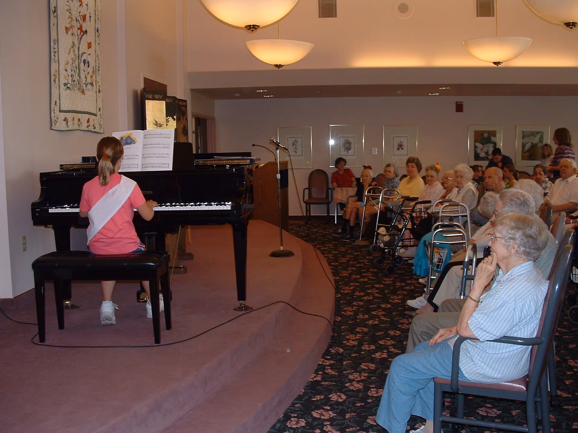 A young girl in a pink shirt and white sash is playing a grand piano on a small stage in front of an audience of elderly people seated in chairs and using walkers in a room with floral carpet and framed artwork on the walls.