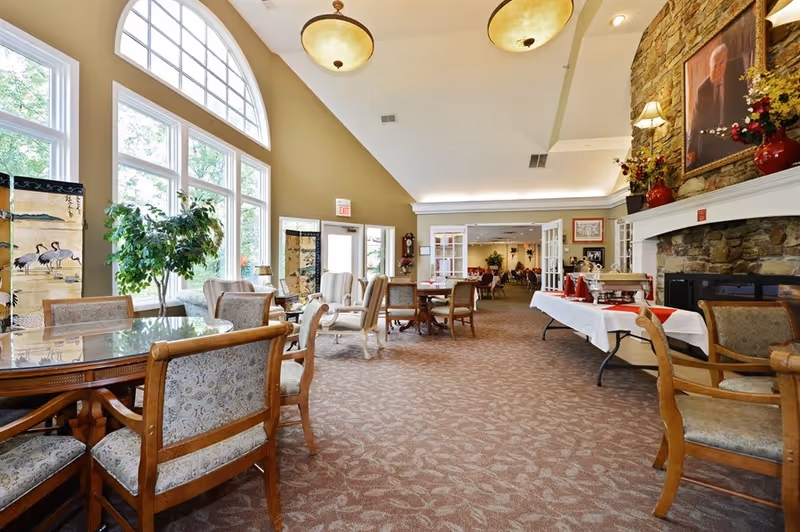 Spacious senior living dining/lounge area with tables and chairs, large arched windows, and a buffet table by a stone fireplace.