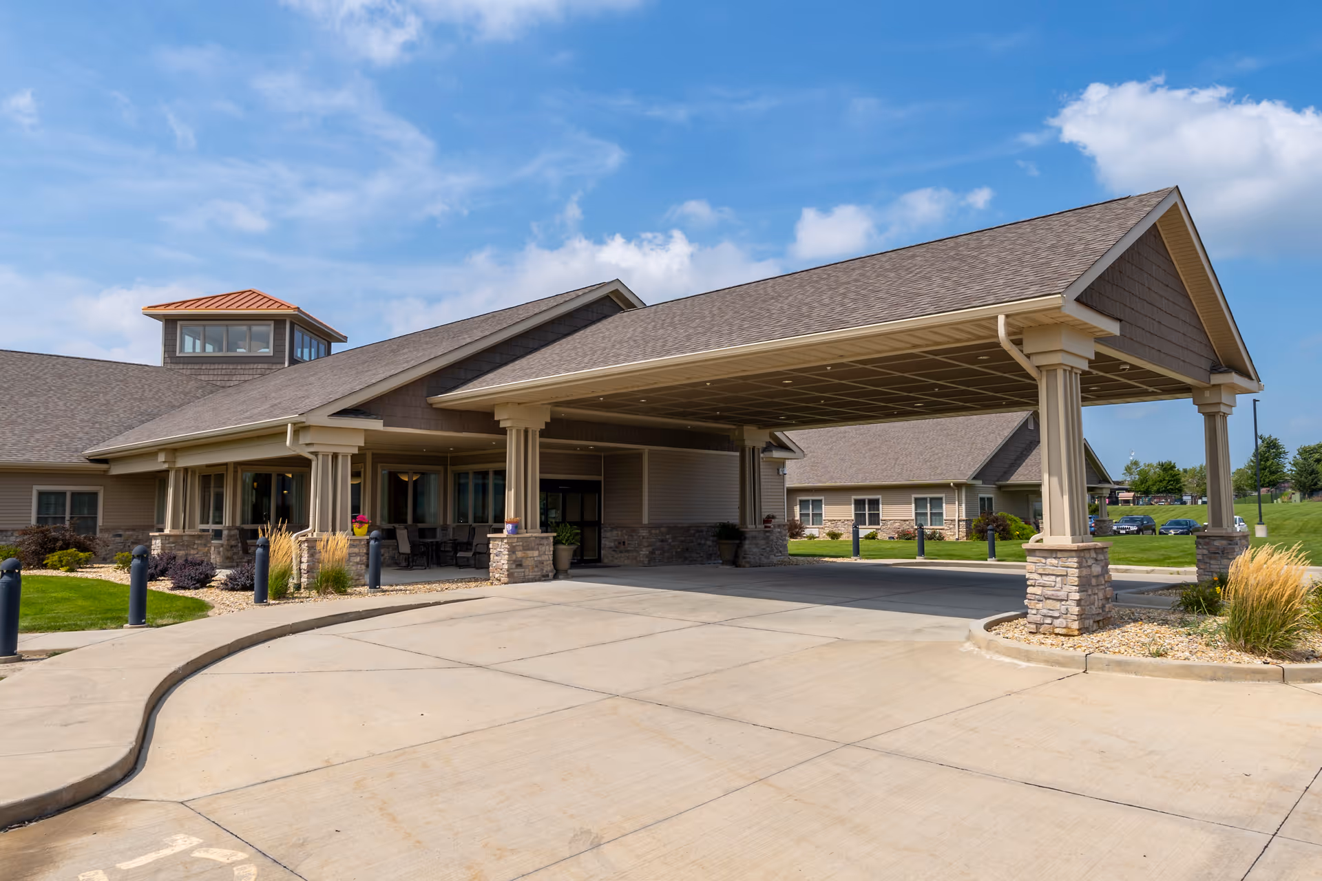 Exterior view of The Carriages of Bloomington senior living facility showing a covered entrance with stone pillars, beige siding, and a well-maintained driveway under a partly cloudy blue sky.