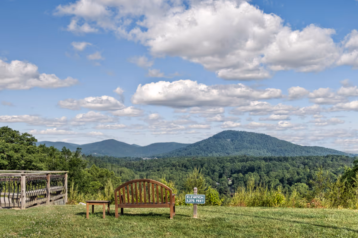A scenic outdoor view from Heather Glen At Ardenwoods showing a wooden bench and small table on a grassy area overlooking a forested valley and distant mountains under a partly cloudy blue sky. A small sign near the bench indicates the elevation as 2,275 feet.