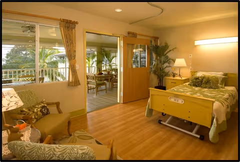 A cozy bedroom in a senior living facility with a single bed featuring a green and white floral bedspread. The room has wooden flooring, a bedside table with a lamp, and a potted plant in the corner. There are two armchairs with patterned cushions and a small table with a lamp. A large window with curtains lets in natural light, and a sliding door opens to an adjacent room with more seating and plants.