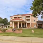 A peach-colored, three-story assisted living facility with a covered entrance and a manicured lawn.