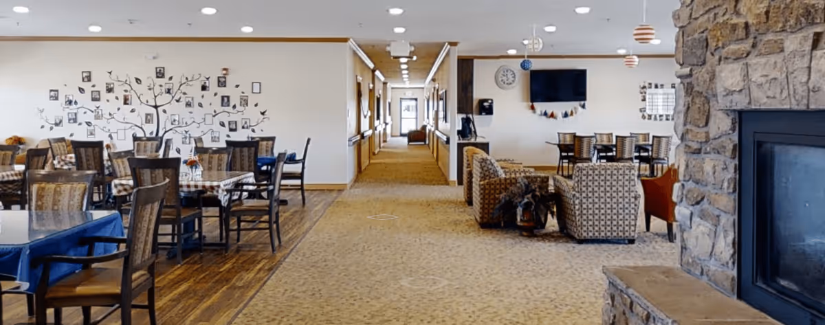 Interior view of a senior living facility showing a dining area with tables and chairs on the left, a hallway in the center, and a sitting area with armchairs and a stone fireplace on the right. The walls are decorated with a tree mural with photos and a clock above a mounted TV.