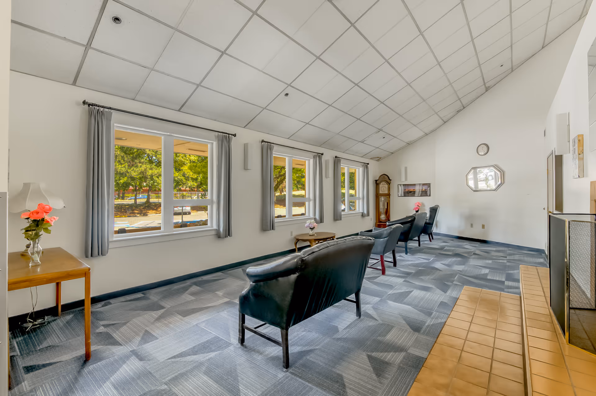 A bright and spacious living room area with large windows letting in natural light. The room features several black leather chairs arranged around small wooden tables with flower vases. There is a grandfather clock in the corner, a wall clock, and a fireplace with a tiled hearth. The walls are white, and the ceiling has a grid pattern with recessed lighting. Outside the windows, trees and a parking lot are visible.