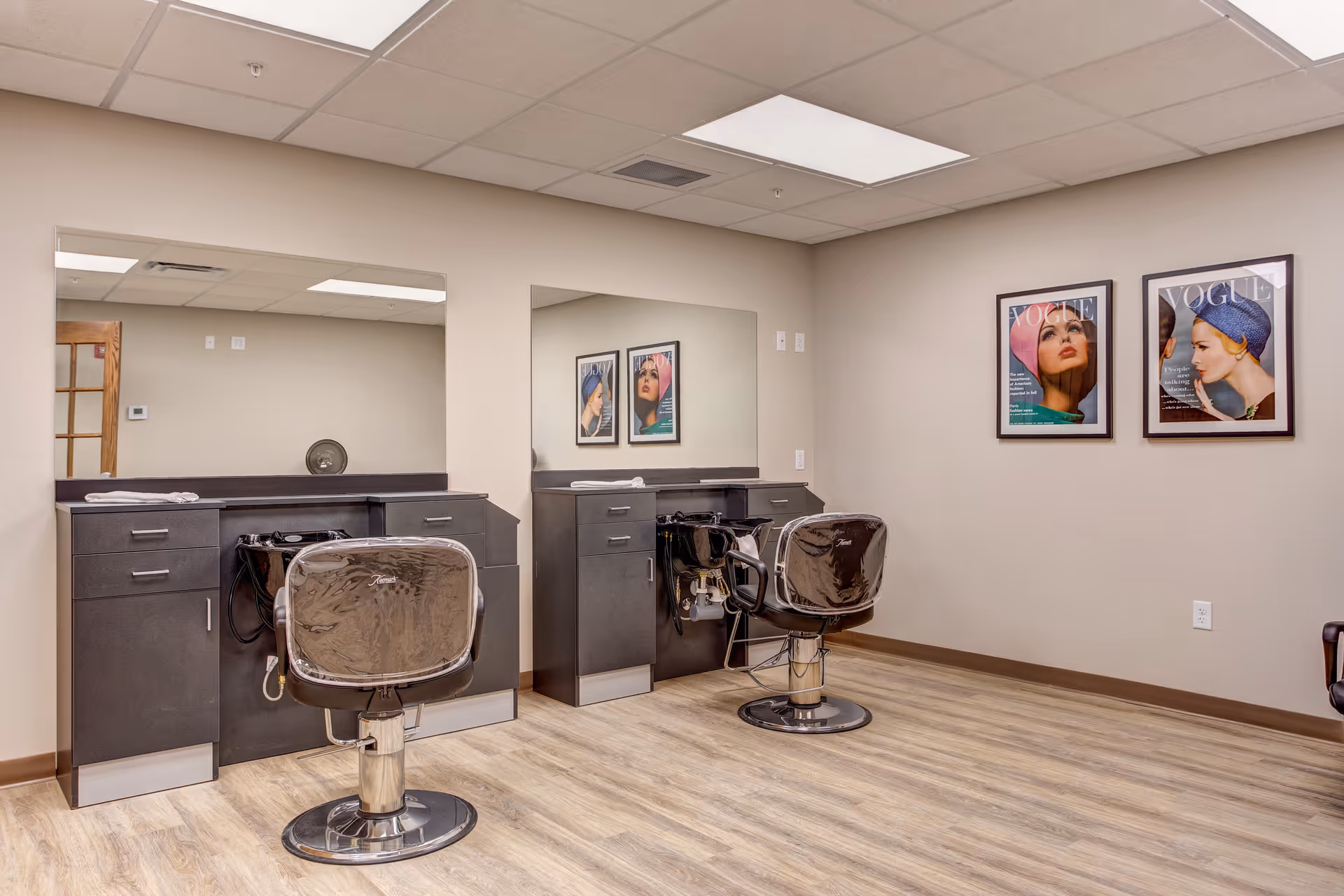 Interior view of a hair salon area in a senior living facility with two black salon chairs in front of two black wash basins and large mirrors. The room has light-colored walls and wood-patterned flooring. Two framed Vogue magazine covers featuring women wearing hats are hung on the wall.