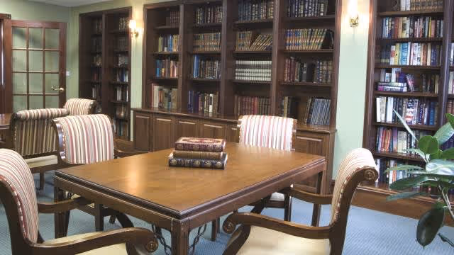 A cozy library room with wooden bookshelves filled with books lining the walls. In the center, there is a wooden table with four upholstered chairs featuring striped fabric. A few books are stacked on the table, and there is a green plant visible on the right side.