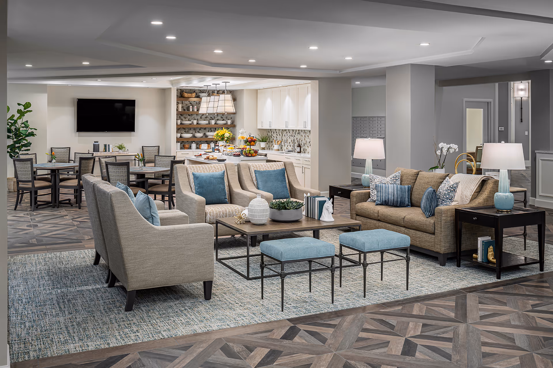 A spacious and modern common area in a senior living facility featuring a seating area with a sofa, armchairs, and ottomans arranged around a coffee table. The room has a patterned rug, wood flooring, and soft lighting from table lamps. In the background, there is a dining area with tables and chairs, a flat-screen TV mounted on the wall, and a kitchen area with white cabinets, open shelves with dishes, and a countertop with fruit and snacks.