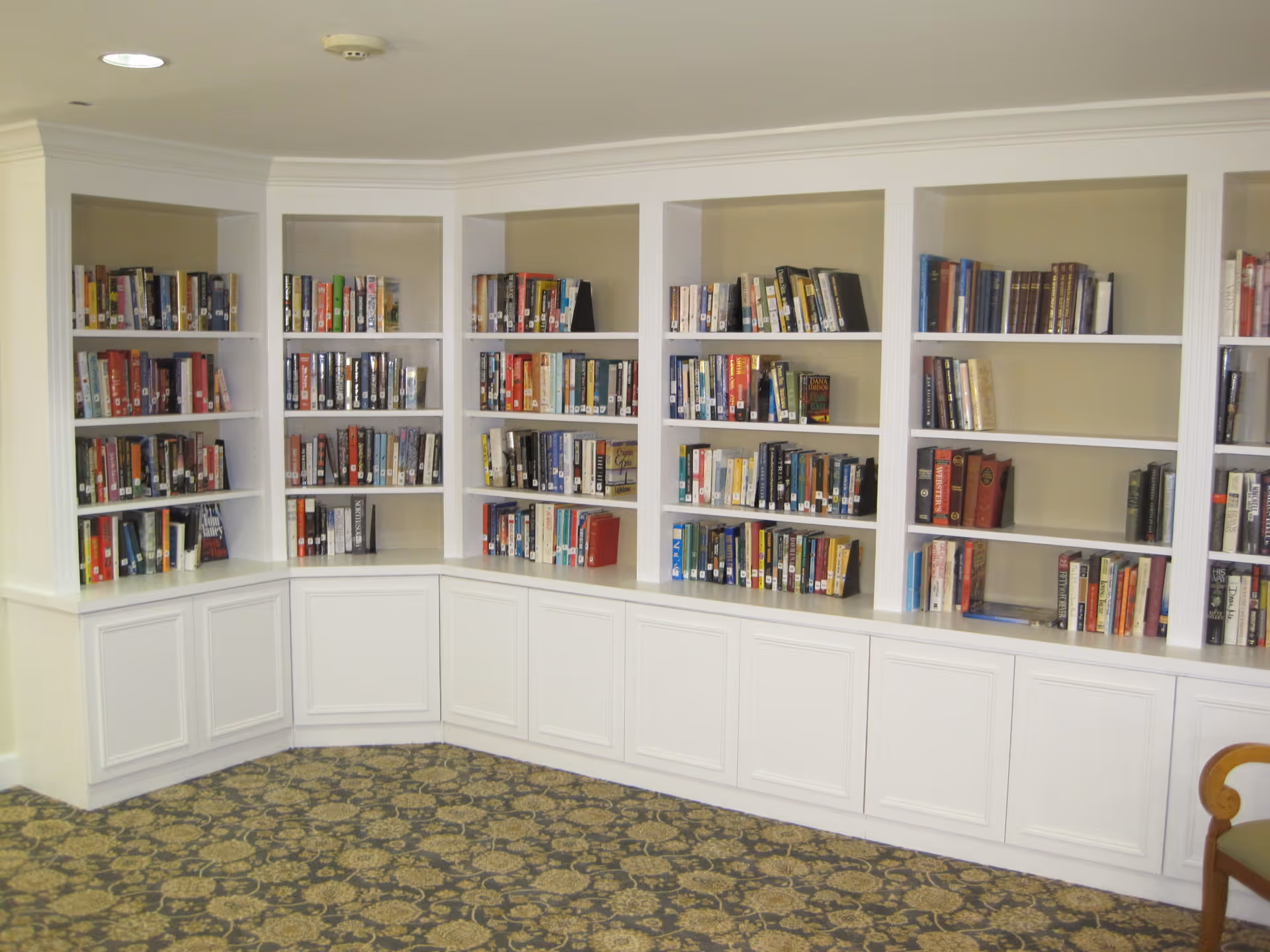 A corner of a room with built-in white bookshelves filled with books. The shelves are set against light-colored walls and above a patterned carpeted floor. A wooden chair with a green cushion is partially visible on the right side.
