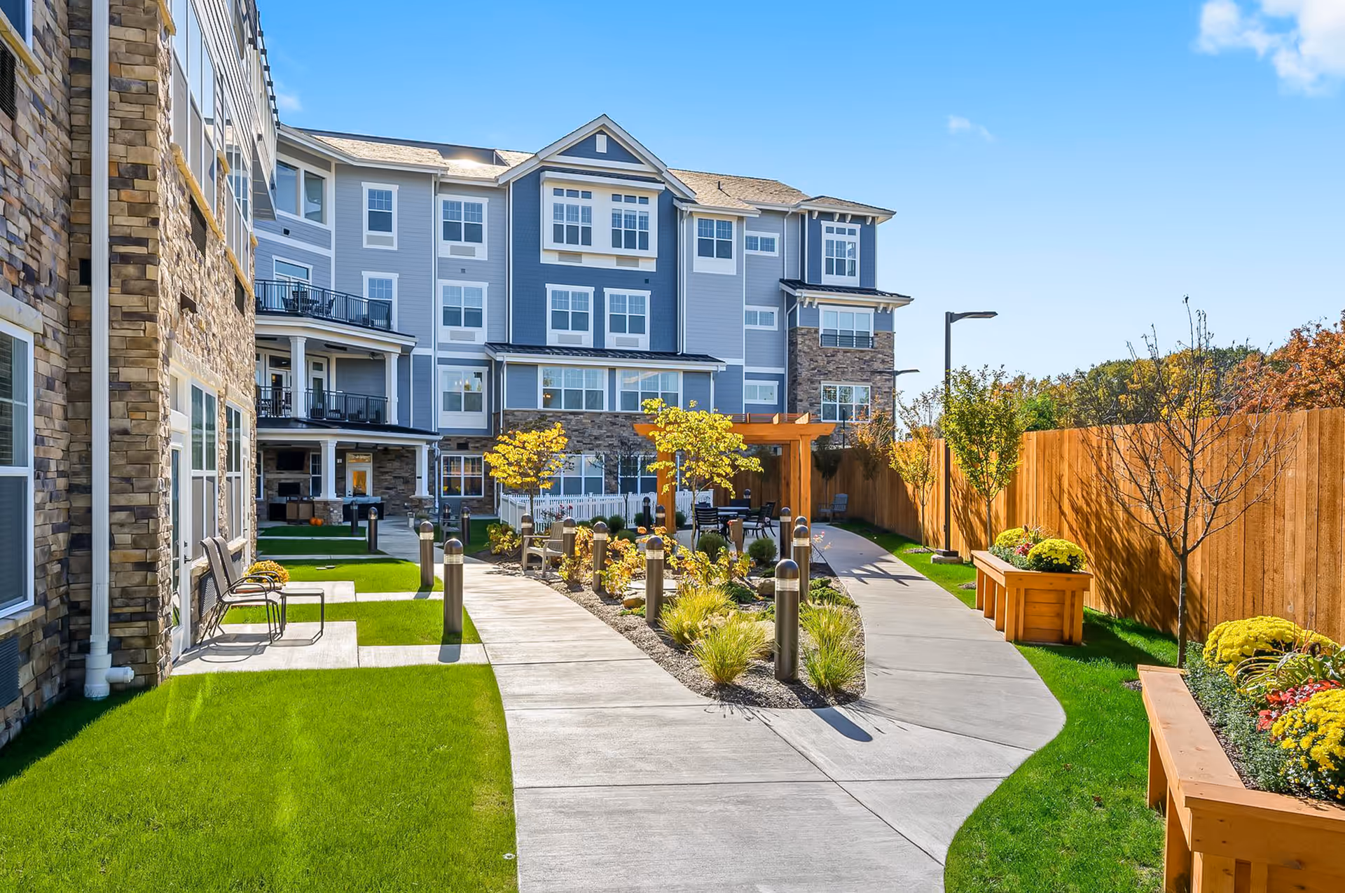 Outdoor courtyard area of The Remington of McCandless featuring a paved walkway, green grass, benches, small trees, flower beds, and a multi-story building with stone and blue siding in the background under a clear blue sky.