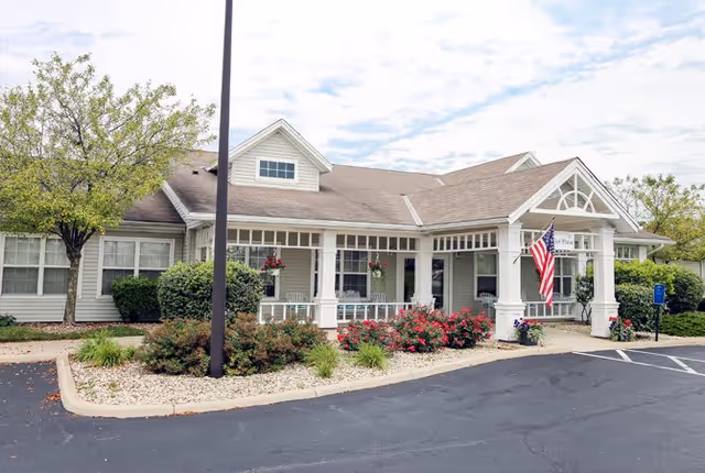 Exterior view of a single-story senior living facility named Taylor Place with a covered entrance, American flag, hanging flower baskets, bushes, and trees. The building has white siding and a gray roof, with a parking lot in the foreground.