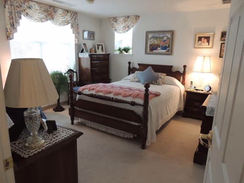 Sunlit bedroom featuring a wooden four-poster bed with bedside tables, lamps, dresser, and floral curtains.