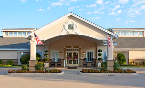 Covered front entrance of a senior living building with a porte-cochere, American flags, rocking chairs, and potted plants.