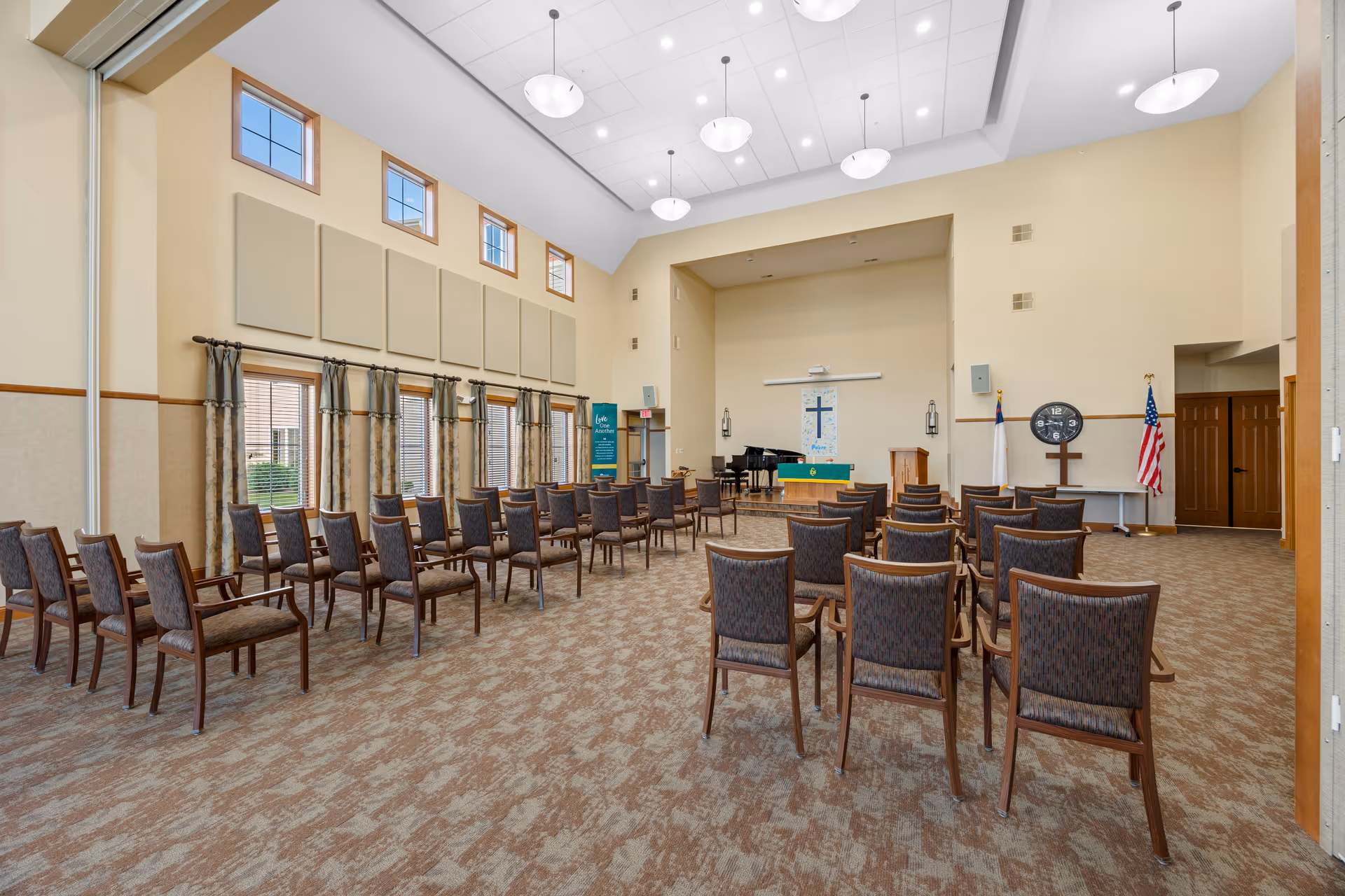 A large, well-lit room with rows of chairs arranged facing a small stage area. The stage has a piano, a podium, and a wall hanging with a cross. There are multiple windows with curtains along one side of the room, and the ceiling has several round light fixtures. An American flag and a Christian flag are positioned near the stage.