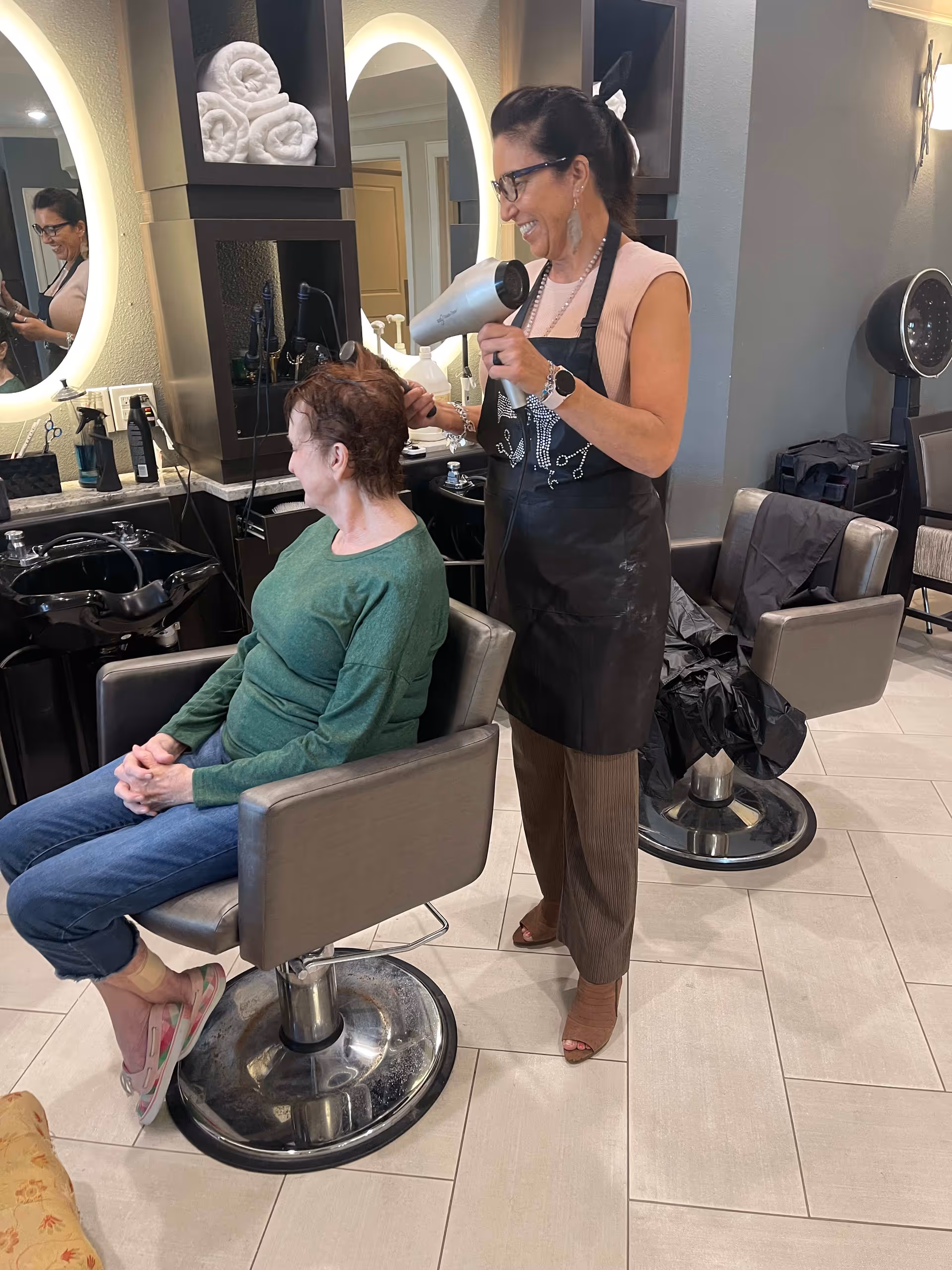 A hairstylist blow-dries a seated woman's hair in a salon area with styling chairs and mirrors.