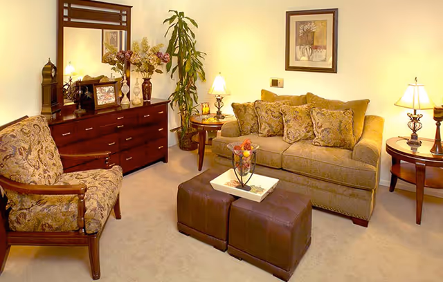 A cozy living room with a beige sofa adorned with patterned cushions, a matching armchair, two brown ottomans serving as a coffee table with a decorative tray and fruit centerpiece, two side tables with lamps, a wooden dresser with a mirror, framed artwork on the wall, and a tall potted plant in the corner.