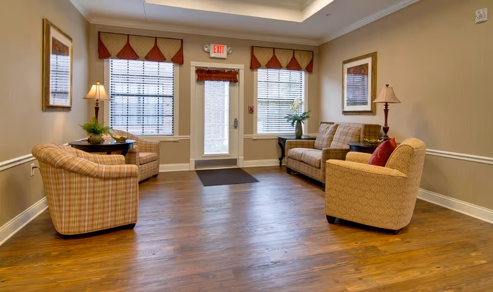 A cozy senior living facility common area with wooden flooring, two armchairs, a sofa, side tables with lamps and plants, framed artwork on the walls, and a glass door with windows on either side letting in natural light.