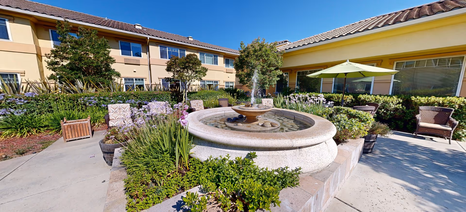 Outdoor courtyard area at Cogir of Sonoma Plaza featuring a circular stone fountain with water flowing, surrounded by lush greenery, flowering plants, and patio seating including chairs and a table with a green umbrella. The building with windows and beige walls is visible in the background under a clear blue sky.