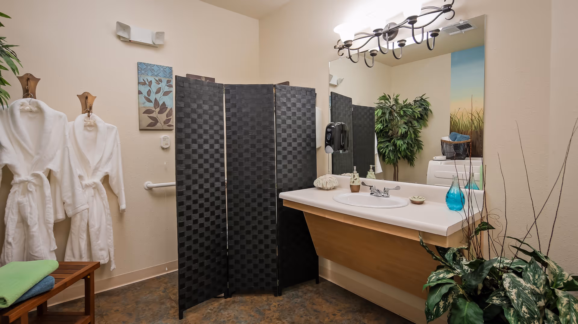 A bathroom area with a large mirror above a sink, a black folding privacy screen, two white bathrobes hanging on the wall, a wooden bench with folded towels, and several plants. The room has beige walls and a decorative light fixture above the mirror.