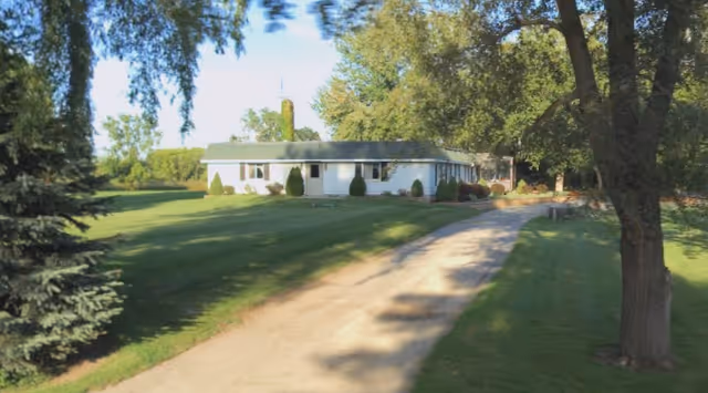 A single-story white building with a green roof surrounded by a large grassy area and several trees. A dirt driveway leads up to the building, which is set in a peaceful, rural environment under a clear blue sky.